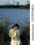 Tranquil Moment by the Lakeside – Woman in White Dress Holding Marigold Flowers with Scenic Water and City Skyline in the Background.