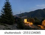 A tranquil mountain cabin at twilight, illuminated by warm indoor lights. Surrounded by tall pine trees and majestic mountains under a starry sky, this cabin evokes a peaceful escape into nature