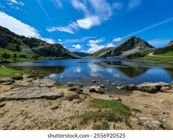 A tranquil mountain lake with clear, calm water, reflecting the blue sky and clouds. Surrounded by rocky hills and lush greenery. The foreground features a rocky and grassy shoreline. - Powered by Shutterstock