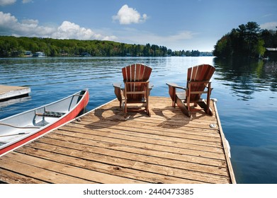 Tranquil Muskoka, Ontario setting as two Adirondack chairs grace a wooden dock by the shimmering waters of a calm lake on a sunlit summer day, accompanied by a canoe and paddles. - Powered by Shutterstock