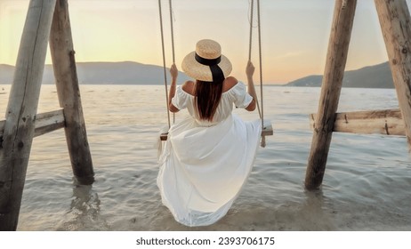Tranquil ocean beach, featuring a young brunette woman in a dress gently swaying on a swing. Her feet caress the sea water, adding a touch of magic to the scene.