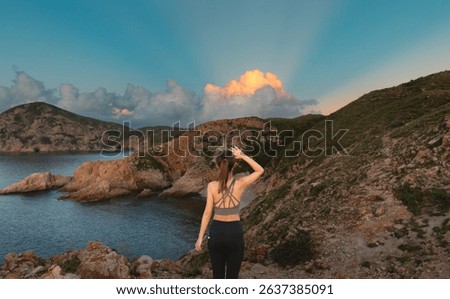 A tranquil panoramic landscape with the back of an Asian girl looking out at layers of dramatic low clouds catching the rays of orange light peering through the coastal hills by the sea at dawn