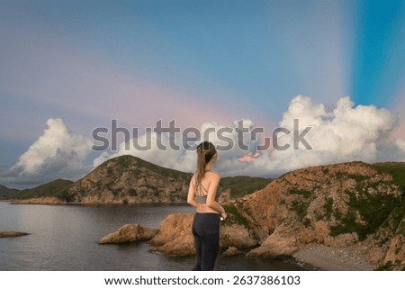 A tranquil panoramic landscape with the back of an Asian girl looking out at the soothing pink morning sky with layers of dramatic low clouds peering behind the coastal hills by the sea at dawn