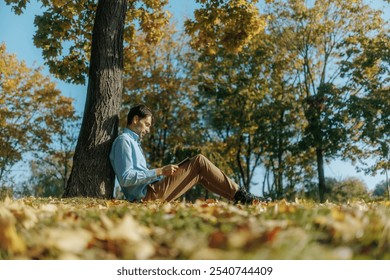 In a tranquil park, a man enjoys a book, sitting on the grass near a tree. The autumn season brings warmth and color to his peaceful reading escape. - Powered by Shutterstock