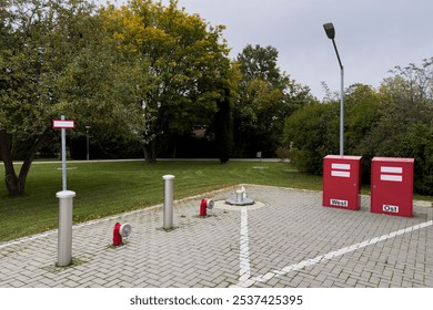 A tranquil park shows two red boxes marked West and Ost and fire hydrants, surrounded by lush greenery and autumn foliage under a cloudy sky. - Powered by Shutterstock