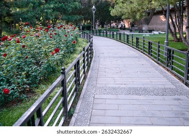 A tranquil paved path through a park lined with red flowers, trees, and green grass, offering a calming natural environment for leisurely walks.