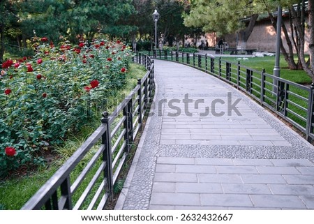 A tranquil paved path through a park lined with red flowers, trees, and green grass, offering a calming natural environment for leisurely walks.