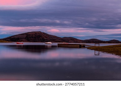 Tranquil Pink Sunset Reflection Over Lake Plastira with Pier and Distant Mountains, Greece.

A wide-angle, long-exposure landscape of Lake Plastira in Thessaly, Greece, captured during the twilight hour. The sky is filled with dramatic, soft clouds tinted with pastel pink and purple hues from the setting sun. The calm, glass-like surface of the lake perfectly mirrors the colorful sky and the rolling silhouettes of the surrounding forested mountains. A wooden pier and a few small boats moored in the distance add a sense of scale and stillness to this peaceful Mediterranean highland scene.