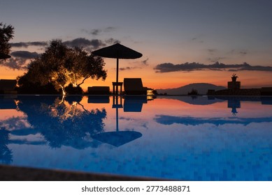 Tranquil resort infinity pool reflecting dramatic sunset sky, with parasol, sunbeds, and warm evening light in a tropical holiday setting.
