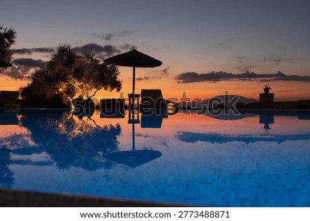 Tranquil resort infinity pool reflecting dramatic sunset sky, with parasol, sunbeds, and warm evening light in a tropical holiday setting.