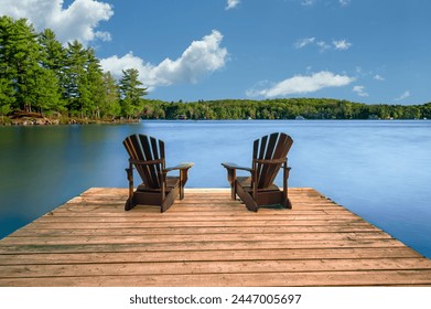 Tranquil Retreat: Two Adirondack chairs on a wooden dock by a serene lake. Rustic cottages nestled among lush trees dot the shoreline. - Powered by Shutterstock