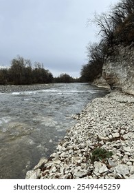 A tranquil river flows between a rocky shore and towering cliffs, surrounded by bare trees under a muted sky. A serene scene showcasing the raw beauty of nature