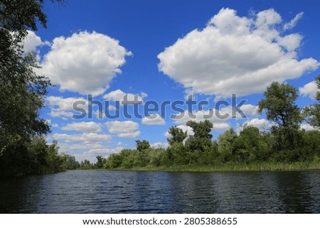 A tranquil river flows gently between verdant, tree-lined shores, with a vast expanse of blue sky dotted with cumulus clouds overhead