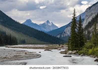 A tranquil river flows gracefully through a secluded valley, embraced by towering mountains and dense forests under a cloudy sky. Nature's beauty is displayed in every direction.