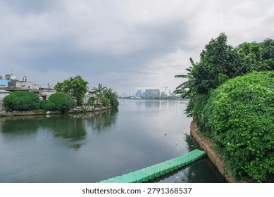 A tranquil river flows through a vibrant landscape, bordered by dense green foliage and a distant urban skyline under an overcast sky.