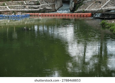 A tranquil river scene featuring reflections of trees and a structure in the background. The water is calm, creating a mirror-like effect. The image captures the beauty of nature.