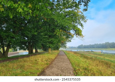 Tranquil riverside walkway bordered by dense green trees and grass on a sunny day. the meandering path invites leisurely strolls, with a river gently flowing nearby, creating an idyllic natural scene. - Powered by Shutterstock