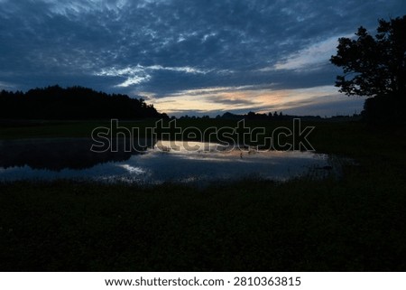 A tranquil rural pond reflecting dramatic pre-dawn clouds and golden sky tones at sunrise in the Selija region of Latvia, with silhouetted trees and reed-lined shores.