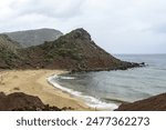 A tranquil scene of Cala Pilar in Menorca featuring a secluded beach with golden sand and a rugged rocky hill under an overcast sky.