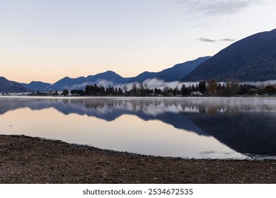 A tranquil scene of a calm lake reflecting nearby mountains and a peaceful landscape during dusk in Mission, BC, Canada. The serene atmosphere and reflections create a sense of tranquility. - Powered by Shutterstock