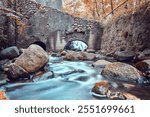 A tranquil scene of an old water mill, nestled amidst autumn foliage. The rushing water flows beneath the stone archway, creating a peaceful and picturesque setting.