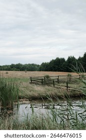 A tranquil scene showcasing an expansive open field, a rustic wooden fence, and a gentle waterway beneath a cloudy sky