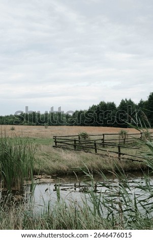 A tranquil scene showcasing an expansive open field, a rustic wooden fence, and a gentle waterway beneath a cloudy sky