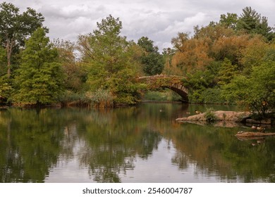 A tranquil scene of a stone bridge over a reflective pond, surrounded by lush greenery and calm waters under a cloudy sky, capturing the serene beauty of nature in the fall. - Powered by Shutterstock