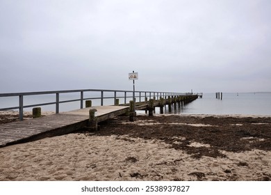 A tranquil scene of a wooden pier reaching into still ocean waters under a cloudy sky. The sandy beach leads to a peaceful horizon, conveying solitude and serenity. - Powered by Shutterstock