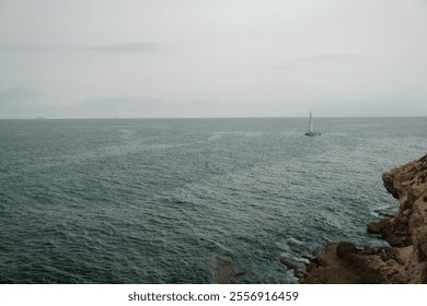 Tranquil seascape with distant sailboat, rocky shore, and moody overcast sky.  themes of serenity, solitude, and maritime exploration. - Powered by Shutterstock