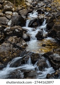 A tranquil stream flows over rocks, surrounded by autumn foliage. The water cascades gently, creating a soothing sound in the peaceful forest setting.