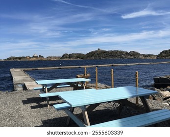 A tranquil summer day in Stavern, Norway, showcasing turquoise picnic benches, a wooden pier, rocky islands, and the blue sea under a clear sky. - Powered by Shutterstock