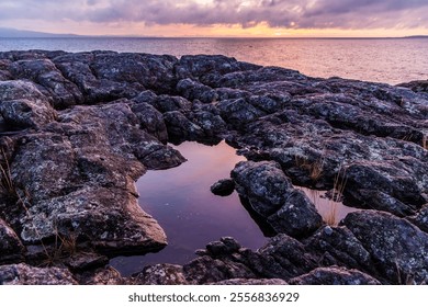 A tranquil sunrise casts a soft glow over the rugged rocky shoreline of Victoria on Vancouver Island, BC. The stunning dawn light reflects in tide pools, creating a picturesque coastal scene. - Powered by Shutterstock