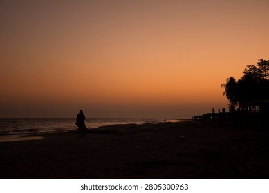 A tranquil sunset beach scene featuring the silhouette of a lone person walking along the shoreline beneath a glowing orange sky. The calm sea, fading daylight, and dark coastal silhouettes create a peaceful atmosphere, capturing the beauty and serenity of the evening by the water.
