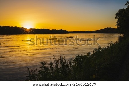 Tranquil sunset on Missouri River; trees and plants in foreground; forest in background; setting sun reflects in water