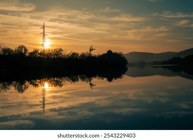 A tranquil sunset scene featuring the silhouette of a power line tower reflected on a calm river, framed by trees and mountains in the distance. - Powered by Shutterstock