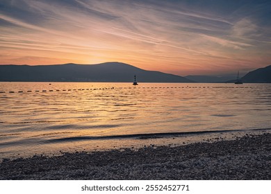 Tranquil sunset scene over calm water with buoys, a small lighthouse, sailboat, and mountains in the background, reflecting the golden light of the setting sun - Powered by Shutterstock