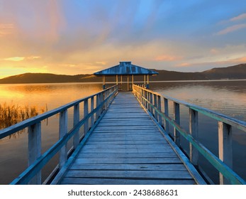 A tranquil sunset scene with a wooden pier leading to a gazebo on a calm lake, framed by distant mountains - Powered by Shutterstock
