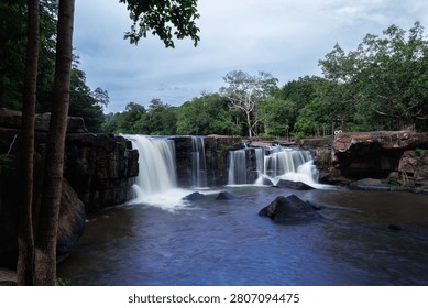 A tranquil view of the multitiered river waterfall flowing over ancient stone ledges into a pool surrounded by lush tropical vegetation at tad ton waterfall.