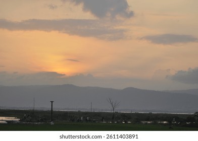 A tranquil view of the sun setting behind distant hills, with a calm landscape featuring scattered trees under a luminous orange sky, evoking feelings of serenity and the beauty of nature.