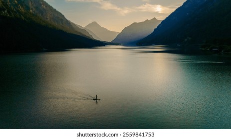 The tranquil waters of Plansee reflect the surrounding mountains, bathed in the warm glow of golden hour. Nature showcases its stunning palette, women on sup board