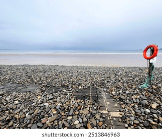 Tranquil Welsh pebble beach with a lifebuoy, calm sea, and overcast sky. Perfect for lovers of coastal minimalism, beach photography, and serene nature landscapes. Ideal for peaceful seascapes. - Powered by Shutterstock