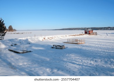 A tranquil winter day in Västanvik, Dalarna, with a blanket of snow covering the landscape. The frozen lake reflects the clear blue sky, while a red cabin and picnic area enhance the peaceful view. - Powered by Shutterstock