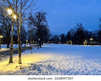 A tranquil winter evening scene in a park, with fresh snow covering the ground and vintage-style street lamps illuminating the pathway. Bare trees line the snow-covered path under a deep blue twilight