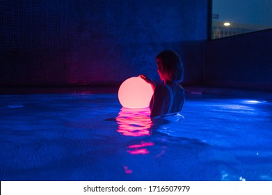 Tranquil young female in swimsuit holding luminous ball while standing in dark swimming pool with neon light