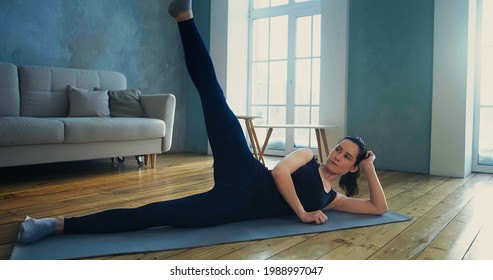 Tranquil Young Woman In Stylish Black Tracksuit Does Supine Side Leg Raises On Mat Against Sofa In Living Room