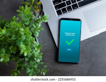 Transaction completed notification in a mobile phone screen, phone alongside with potted plant and laptop on dark stone background