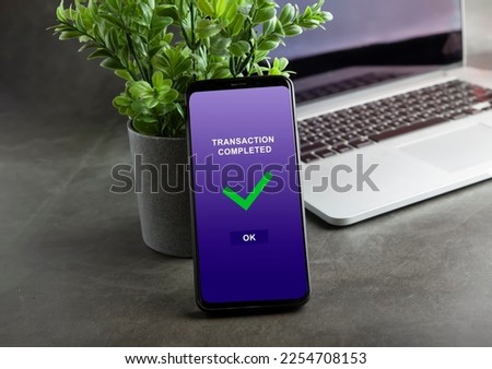 Transaction completed notification in a mobile phone screen, phone alongside with potted plant and laptop