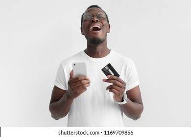 Transaction failed, declined. Young african american man holding credit card and smart phone in hand, crying, yelling because he has no cash, isolated on gray background