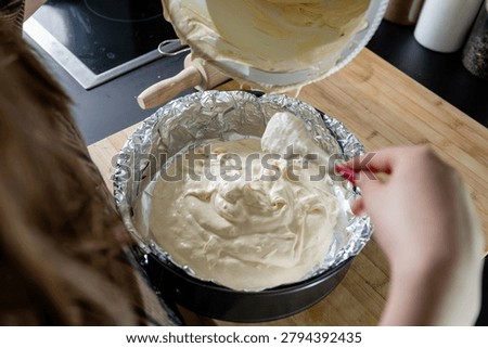 Transferring smooth sponge cake batter from a bowl into a foil-lined springform tin with a red spatula in a modern marble kitchen, preparing a homemade cake.